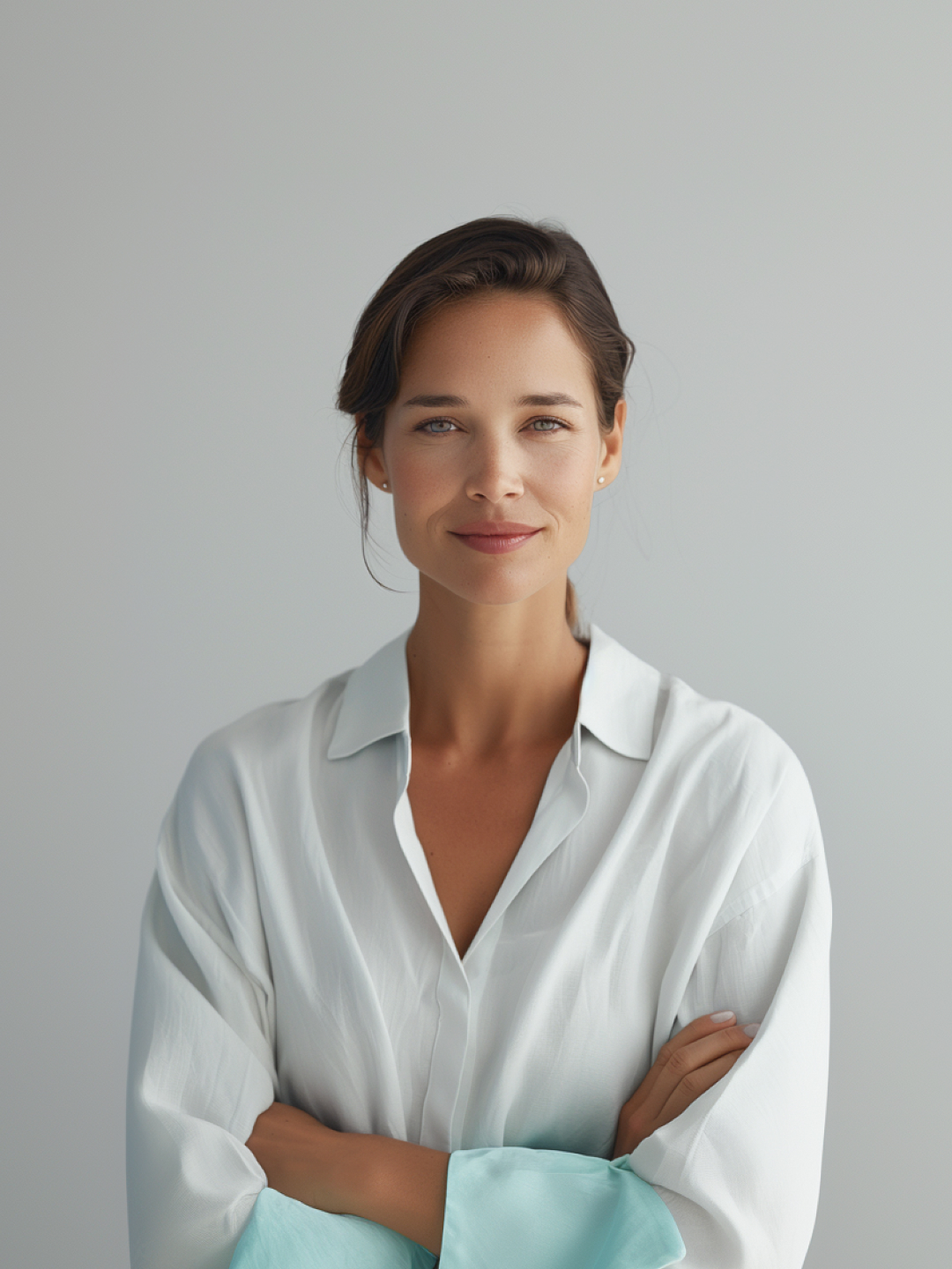 A woman with a confident smile, wearing a white shirt with rolled-up sleeves, stands with her arms crossed against a light gray background.